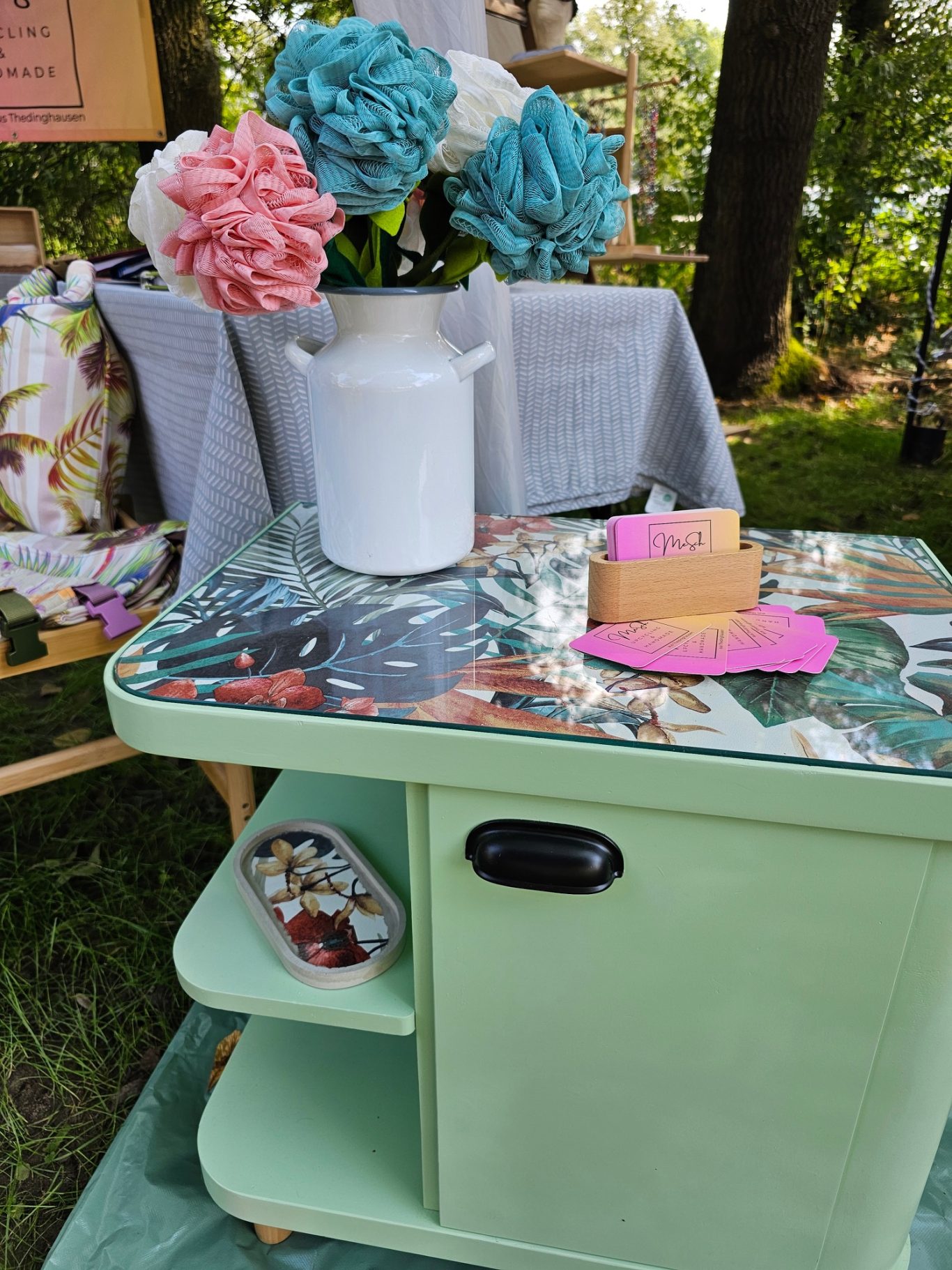 Upgecyceltes Schränkchen Grüner Beistelltisch mit buntem Tischdeckel, Wasserkanne und Blumenarrangement.