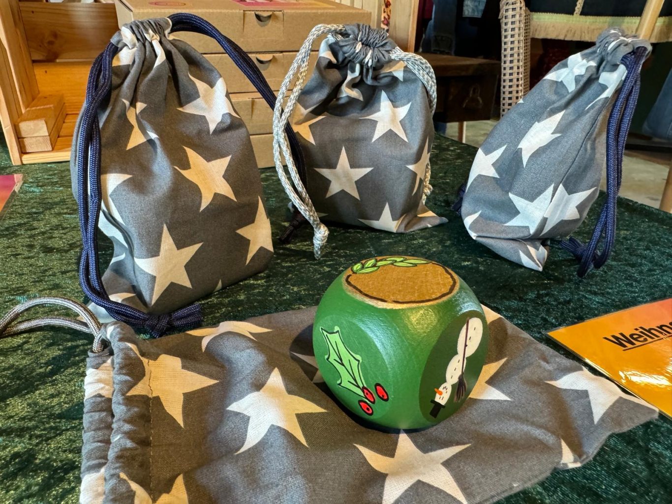 Weihnacht-Geschenke-Würfel Mit Weihnachtsmotiven bemalter Holzwürfel, umgeben von drei grauen Sternenbeuteln.