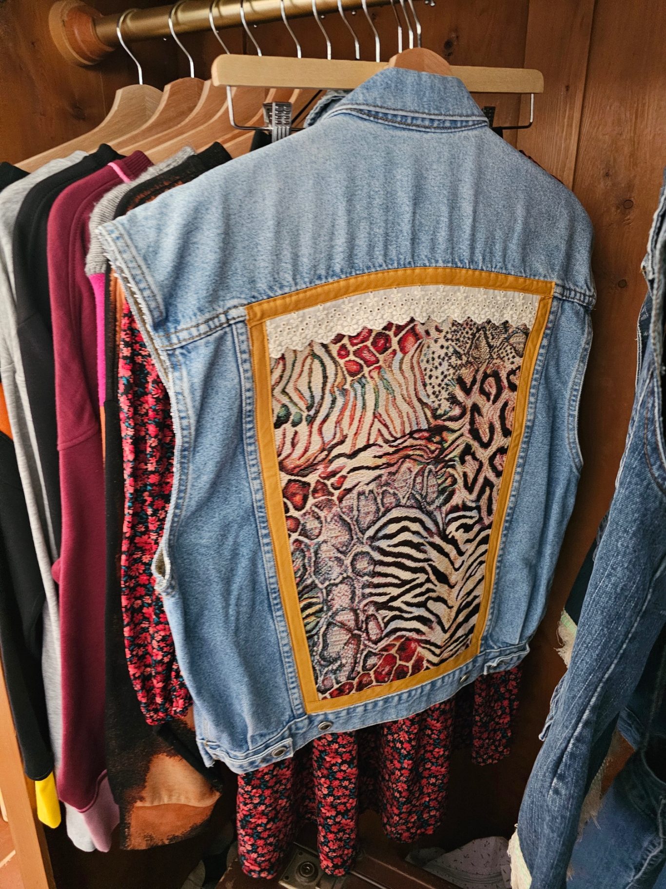 Upgecycelte Tiermuster-Jeansweste Jeansweste mit buntem Tiermuster auf der Rückseite, neben weiteren Kleidungsstücken.
