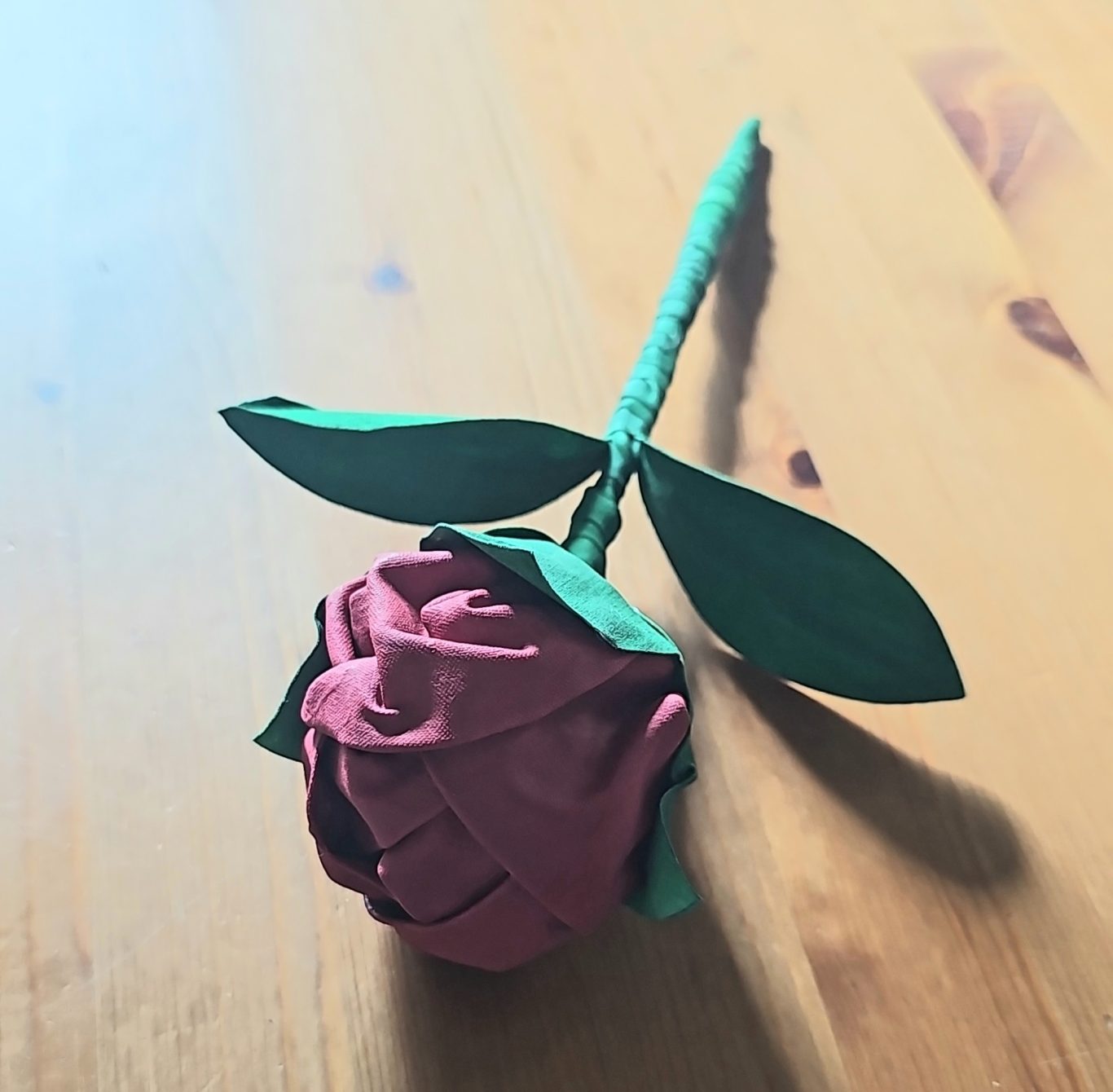 Upgecycelte Rose (Kundenauftrag) Rote Stoffrose mit grünen Blättern auf einem Holzuntergrund.